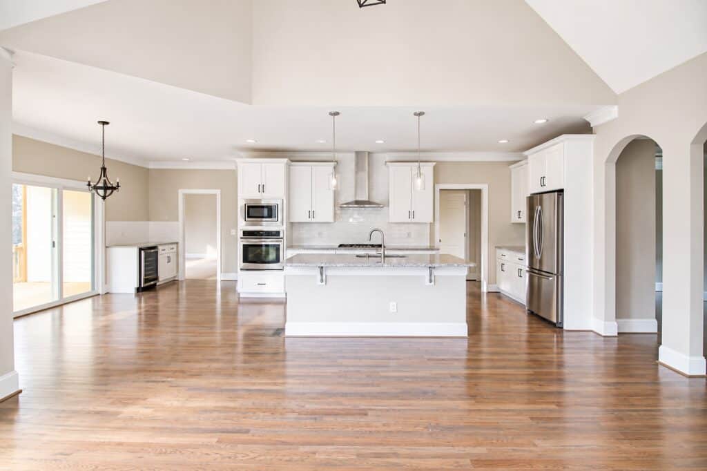 Wide open-concept floor plan displaying a high-end custom kitchen with white shaker cabinets, stainless steel appliances, premium granite countertops, and rich oak hardwood floors running throughout the expansive space.