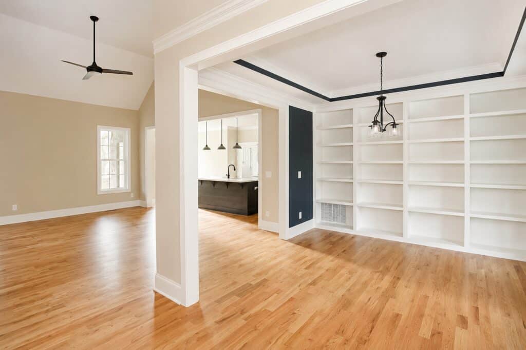 Elegant formal dining room or home library featuring a massive wall of custom built-in white bookshelves, an accented tray ceiling with a modern chandelier, and rich hardwood flooring flowing into the open living area.