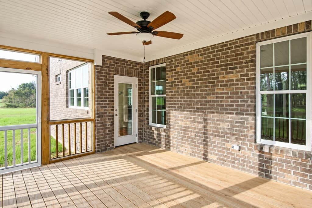 Covered outdoor screened deck area highlighting a rich brown brick veneer exterior wall, double-hung windows, a glass patio door, and low-maintenance wood plank flooring.