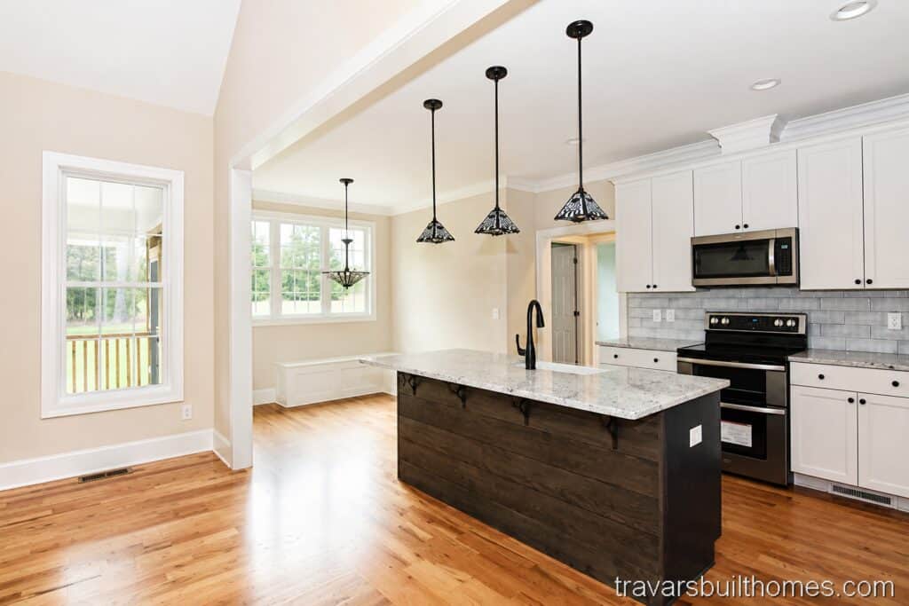 Open view from a custom kitchen island toward a bright breakfast nook with built-in bench seating, highlighting oak hardwood flooring, white shaker cabinets, and industrial cage pendant lights.
