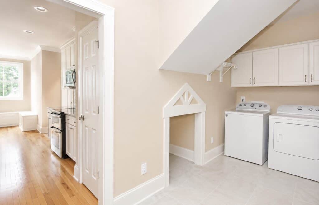 Custom laundry room featuring tile flooring, white cabinetry, a washer and dryer setup, and a unique built-in pet kennel space under the stairs with decorative white gabled trim.
