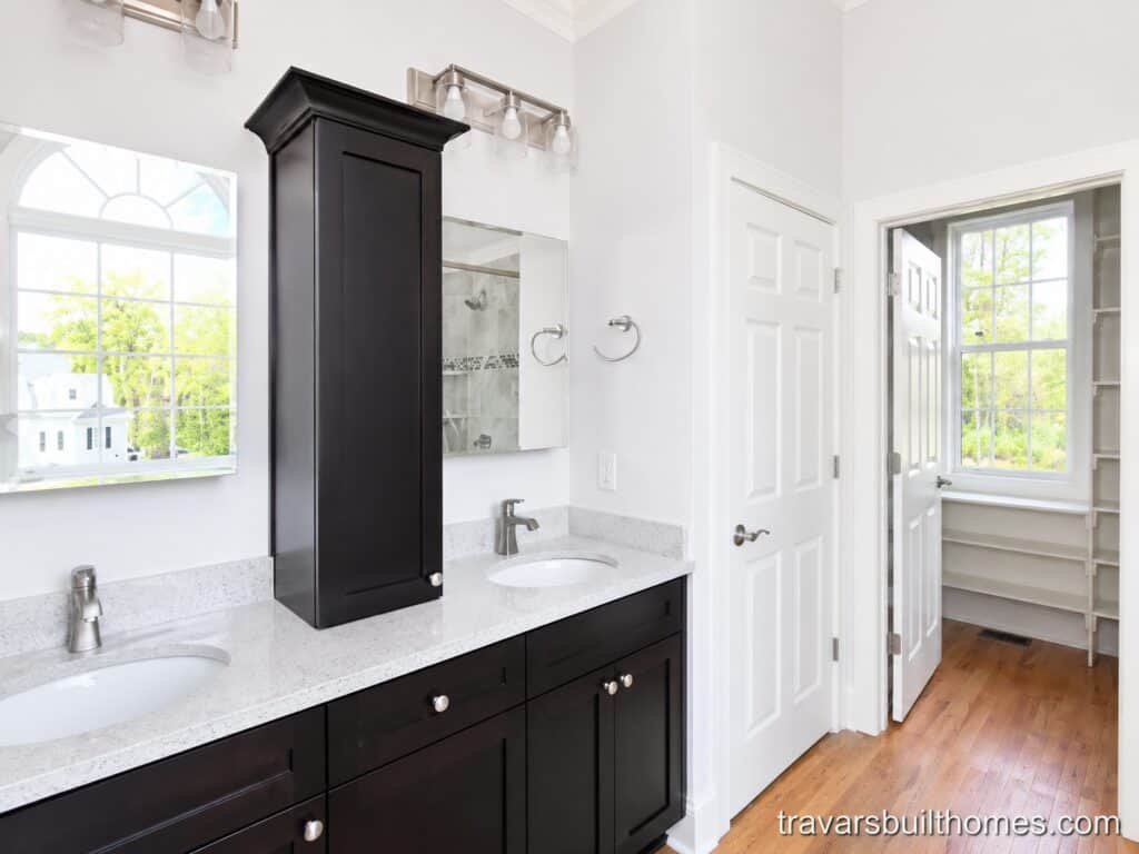Master bathroom double vanity with dark shaker cabinets and white stone countertops, showing an open doorway into a custom walk-in closet with built-in shelving, hardwood floors, and a bright window.