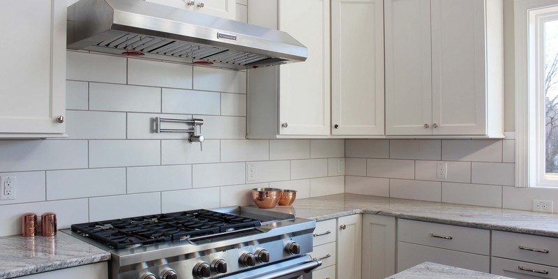 Kitchen range wall with white cabinetry, gray subway tile backsplash, stainless steel range hood, professional-style gas range, granite countertops, corner cabinets, wood flooring, and a bright window beside the counter.