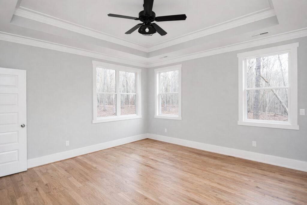 Airy primary bedroom highlighting continuous oak hardwood floors, soft greige walls, three bright windows overlooking the woods, and a multi-tier tray ceiling with a black fan, built by Travars Built Homes.