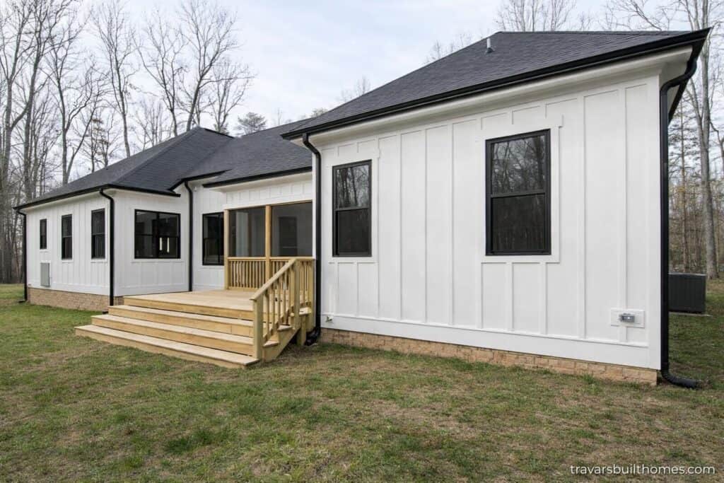 Rear exterior elevation highlighting crisp white vertical siding, black window trim, a brick foundation skirt, and multi-tier wooden deck steps leading down to a grass lawn, built by Travars Built Homes.
