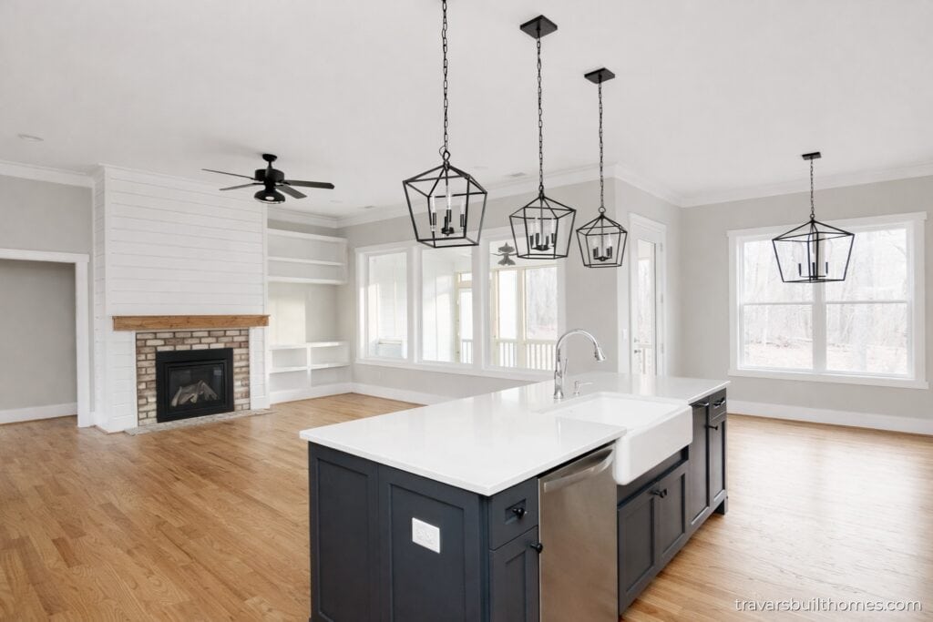 Beautiful open-concept view from behind the kitchen island looking into the great room, showcasing three black geometric lantern pendant lights, a white farmhouse sink, a classic brick fireplace with a wood mantel, and oak floors, built by Travars Built Homes.