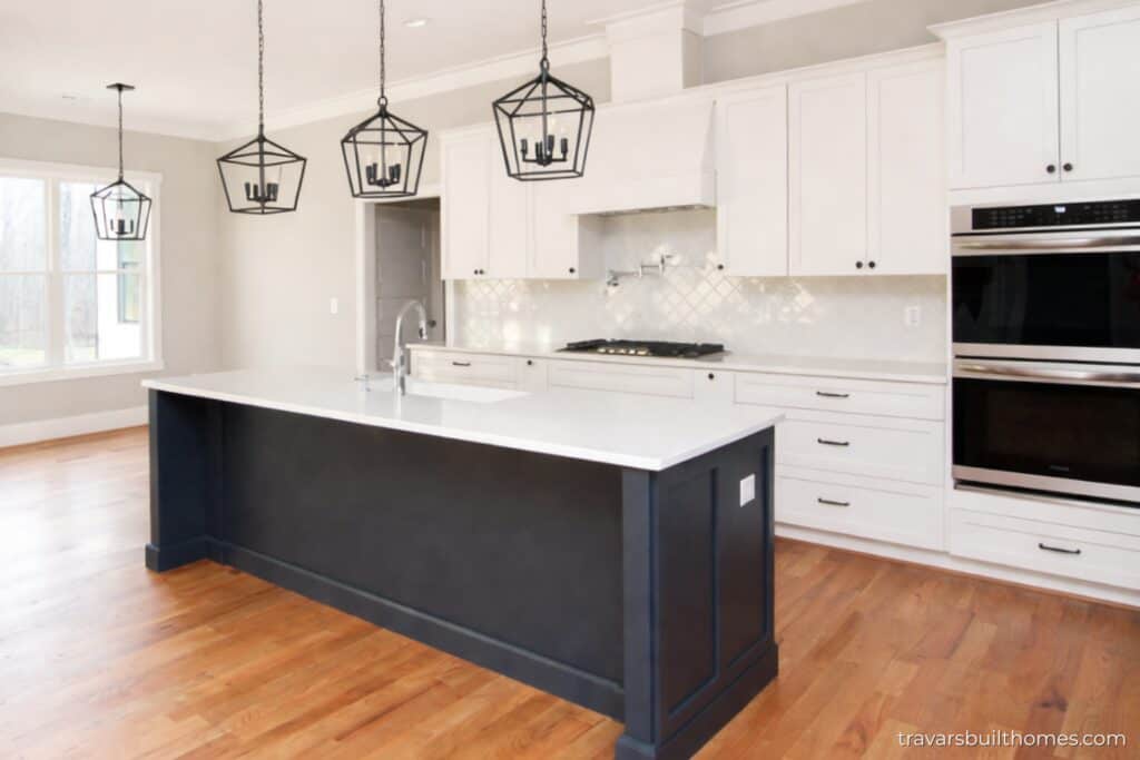 Elegant kitchen profile emphasizing a long dark graphite kitchen island topped with thick white quartz countertops, illuminated by three hanging black iron lantern frame fixtures over oak plank flooring, built by Travars Built Homes.