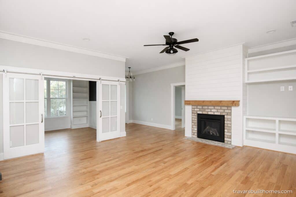 Wide perspective across a light oak great room, featuring a central brick fireplace with a shiplap overmantel alongside double white sliding glass doors that open into a deep navy blue library room, designed by Travars Built Homes.