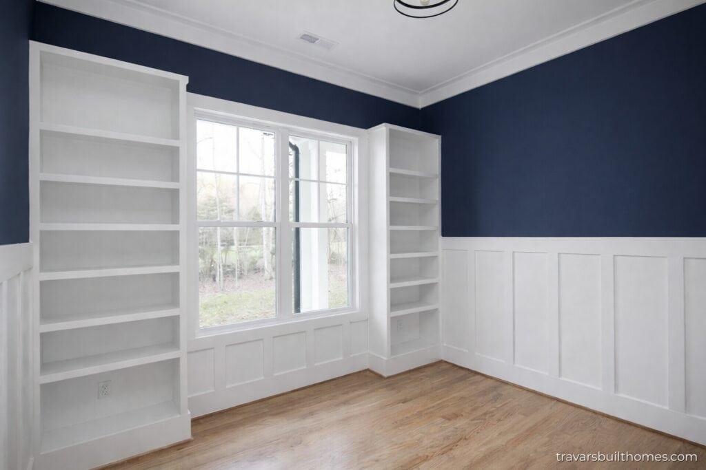 Sophisticated home office space featuring rich oak floors, deep navy blue walls with white wainscoting, and custom built-in white bookshelves framing a wide double window, built by Travars Built Homes.