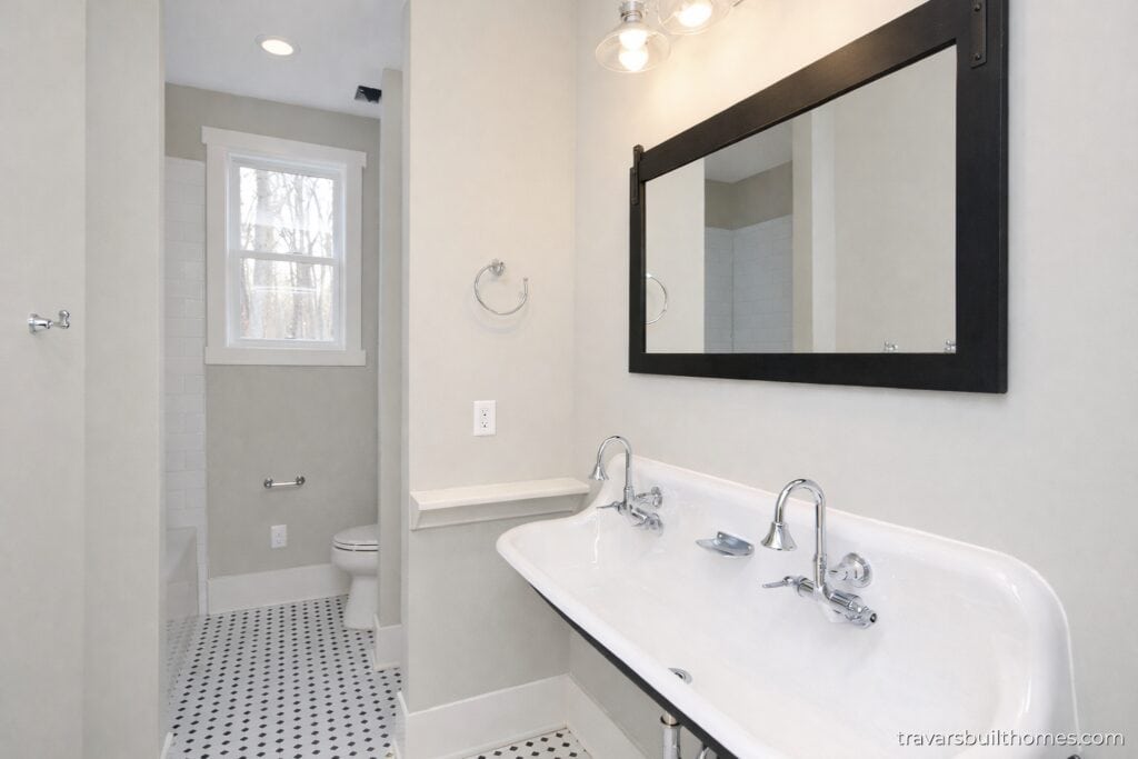 Vintage-inspired guest bathroom featuring a unique double-faucet white porcelain trough sink, a large black-framed mirror, and classic black-and-white mosaic tile floors, designed by Travars Built Homes.