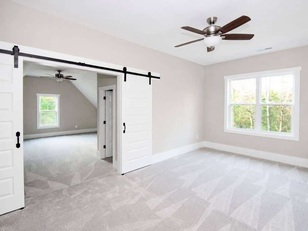 Custom second-floor bonus space detailing an expansive opening with double white sliding barn doors that seamlessly link two large carpeted living areas, built by Travars Built Homes.