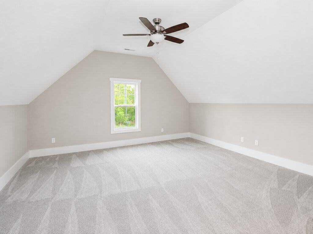 Spacious upstairs bonus room or loft space highlighting angled ceiling lines, a central dark ceiling fan, soft patterned carpeting, and a window overlooking the woods, designed by Travars Built Homes.