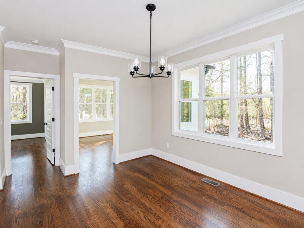 Sunlit dining area or breakfast nook showcasing a modern black chandelier, rich oak floors, and a large triple window providing private views of the surrounding mature forest, designed by Travars Built Homes.