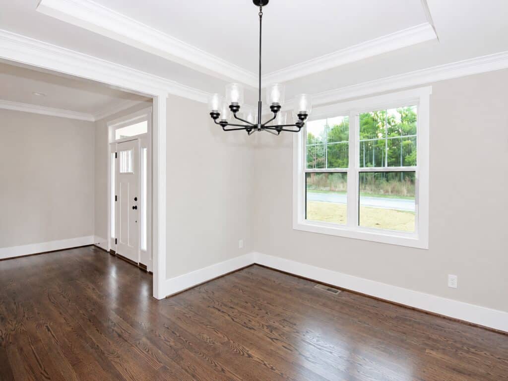 Elegant dining room showcasing a deep tray ceiling with crown molding, a dark iron candelabra chandelier, rich oak flooring, and a large double window overlooking the front yard, by Travars Built Homes.