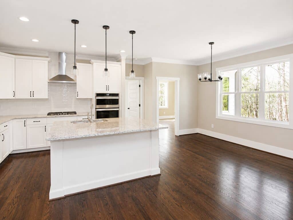 Bright, open custom kitchen showcasing white shaker cabinets, a large island with a speckled granite countertop, a stainless steel chimney hood, and dark oak hardwood floors, designed by Travars Built Homes.
