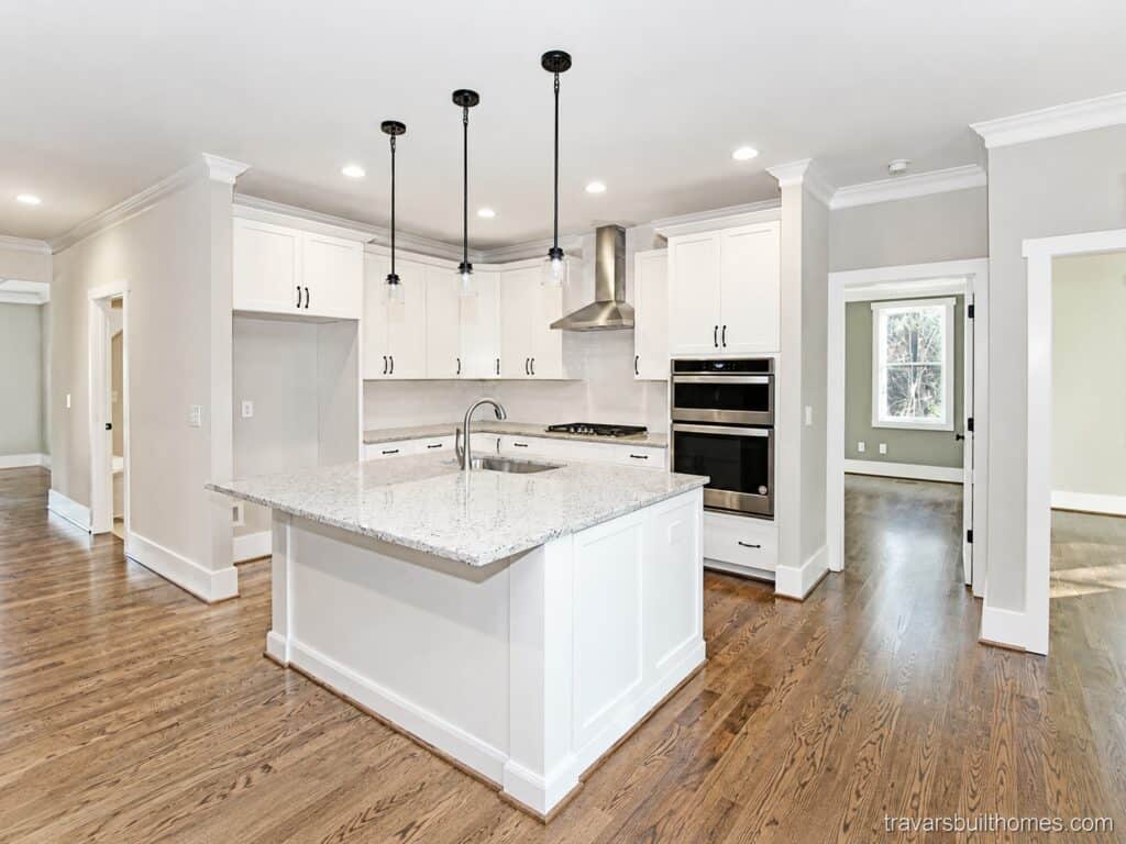 Custom kitchen layout featuring a large central island, crisp white perimeter cabinetry, a stainless steel chimney range hood, and integrated double wall ovens, built by Travars Built Homes.