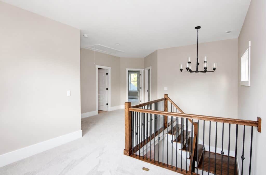 Open second-story loft and landing area showcasing a modern multi-light chandelier, plush carpeting, and a protective wooden handrail with black iron balusters over the stairs, crafted by Travars Built Homes.