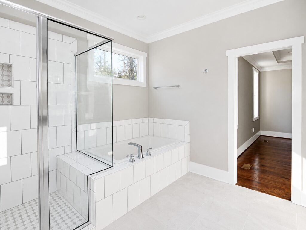 Alternative view of the primary bathroom highlighting a tiled drop-in soaking tub beneath a window, a glass shower enclosure with a mosaic tile floor, and a doorway leading to a hardwood hallway, designed by Travars Built Homes.