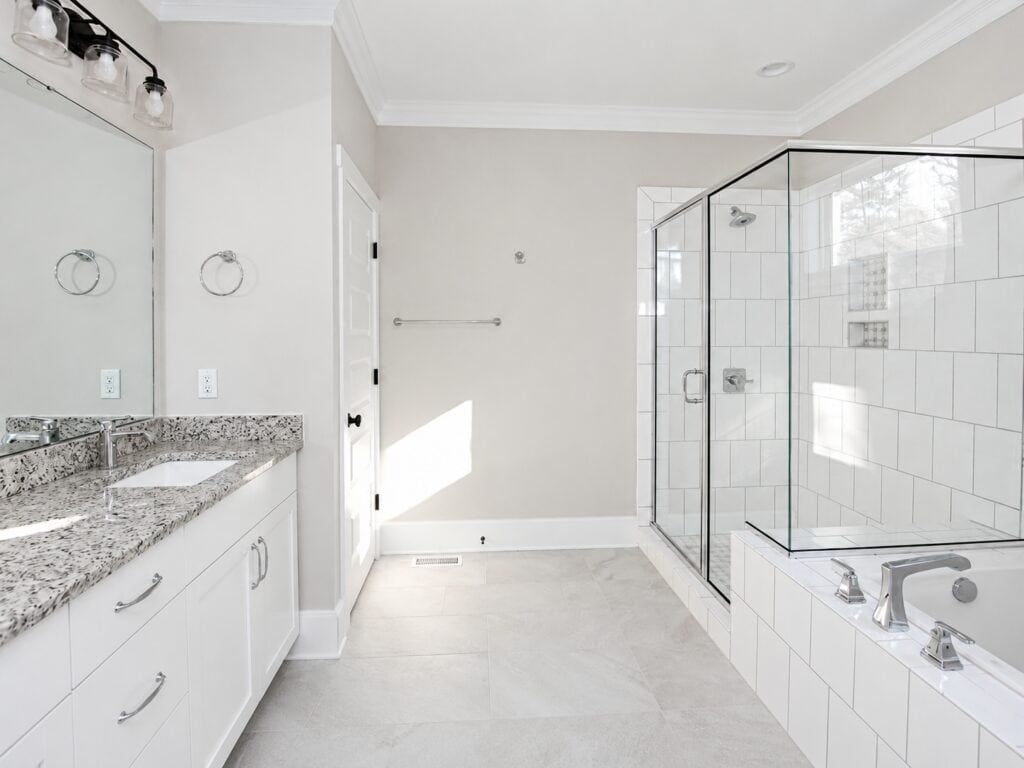 Primary en-suite bathroom featuring a long white vanity with a speckled granite countertop, contemporary black light fixtures, a glass-enclosed shower with white subway tile, and a built-in tub, built by Travars Built Homes.