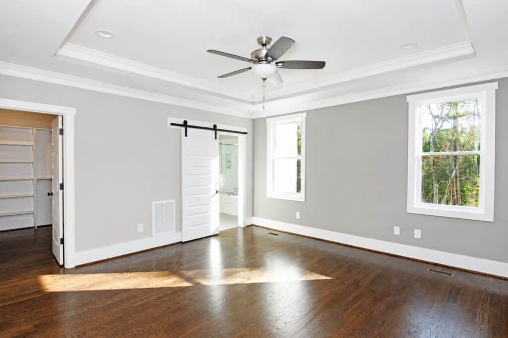Grand primary bedroom suite highlighting a deep tray ceiling with a central ceiling fan, rich wood floors, a dual window layout, and white sliding double barn doors opening to the en-suite, built by Travars Built Homes.
