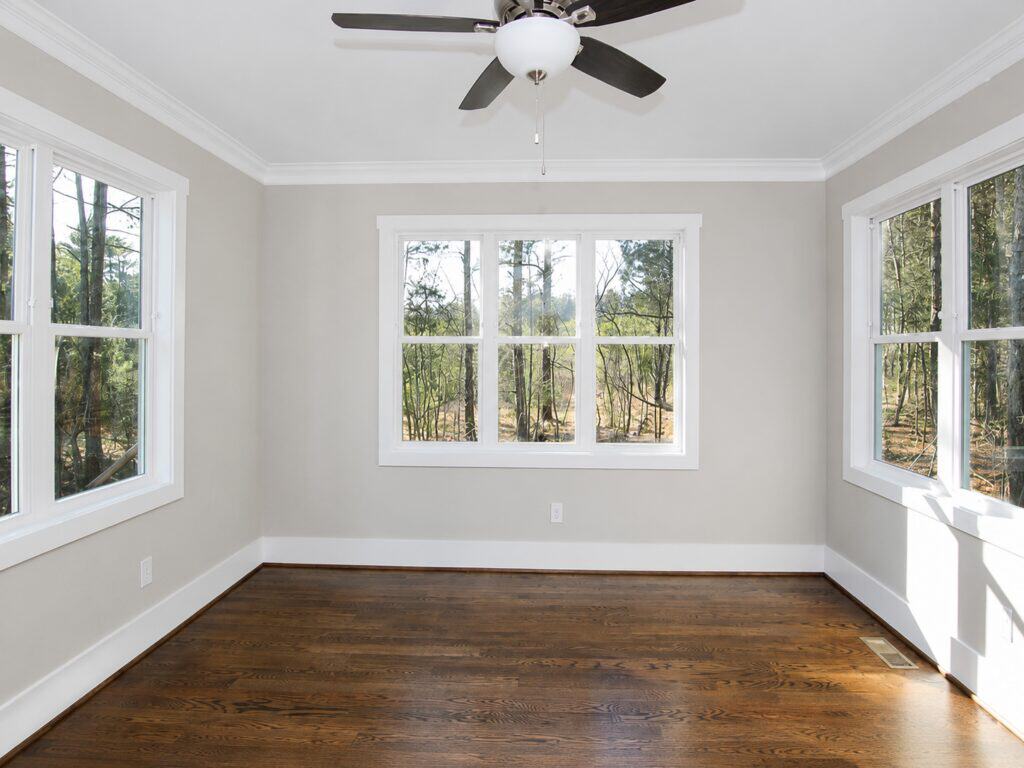 Dedicated home office or study area featuring rich dark oak flooring, a classic multi-blade ceiling fan, and large windows on two walls providing views of the surrounding woods, crafted by Travars Built Homes.