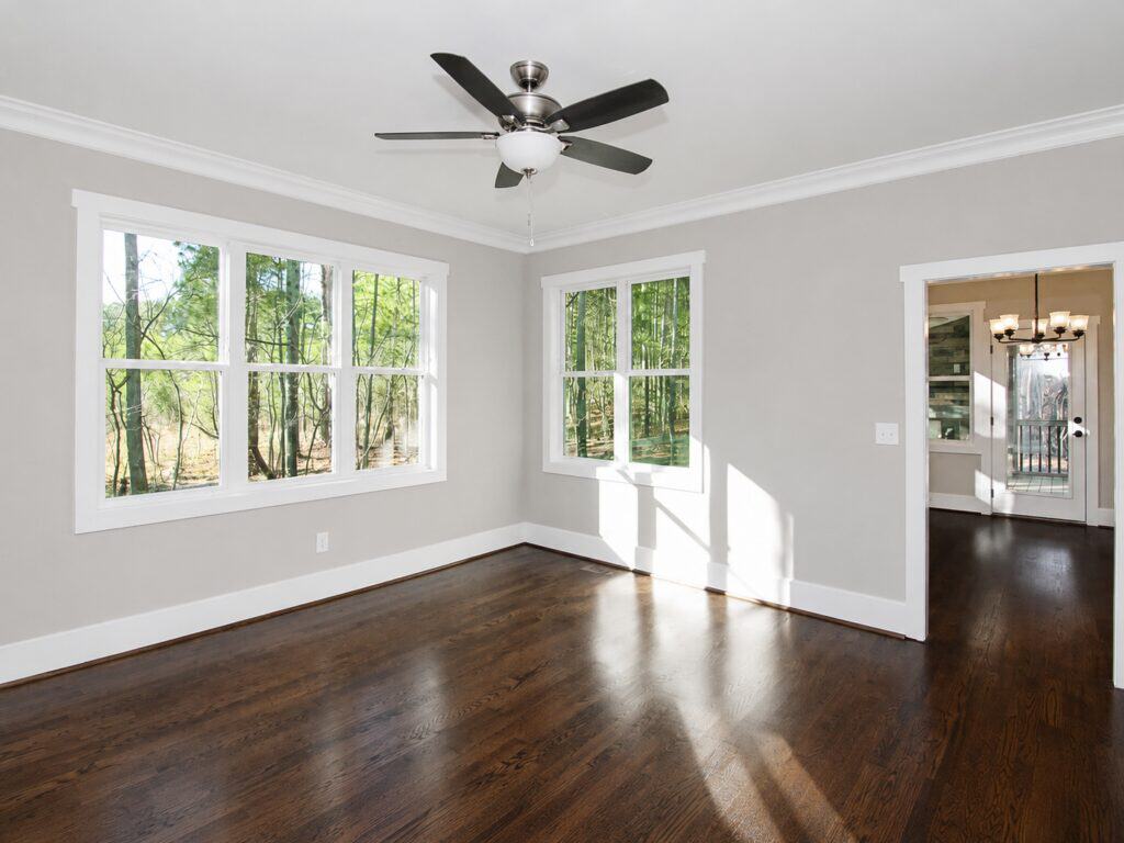 Bright home office or flex room featuring rich dark hardwood flooring, a multi-blade ceiling fan, and large double windows highlighting serene wooded views, designed by Travars Built Homes.