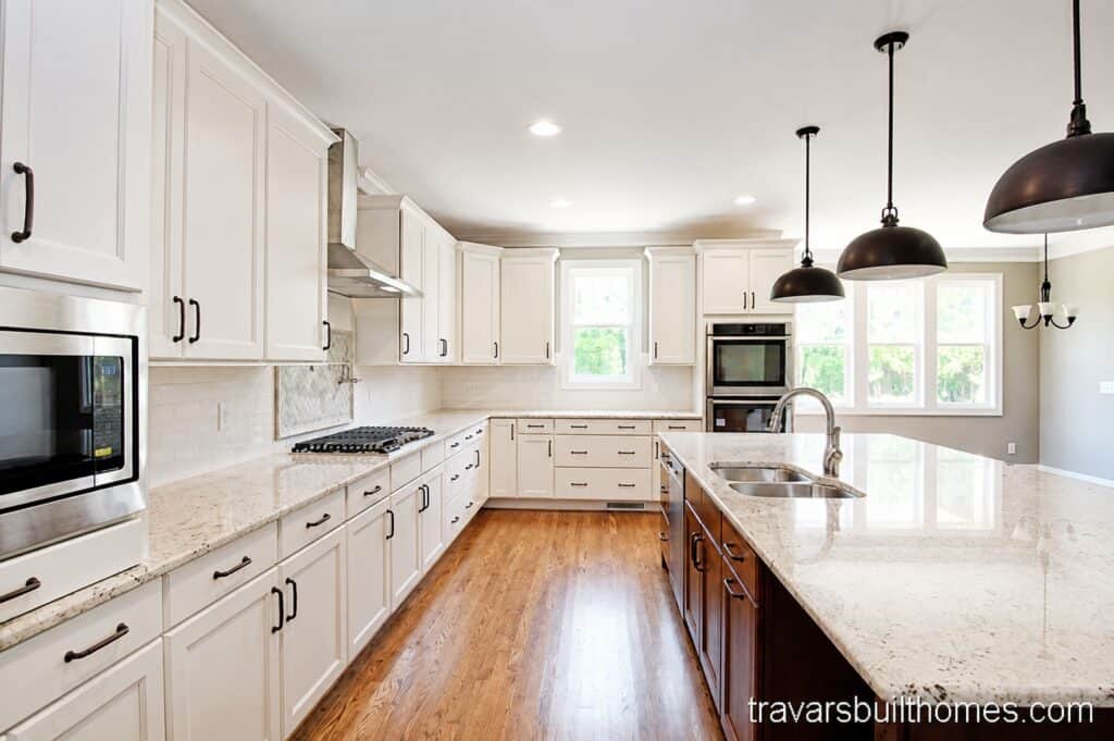 Bright open kitchen layout featuring continuous oak floors, white shaker perimeter cabinets, dual wall ovens, and a long wood-grained center island with a speckled granite top, designed by Travars Built Homes.