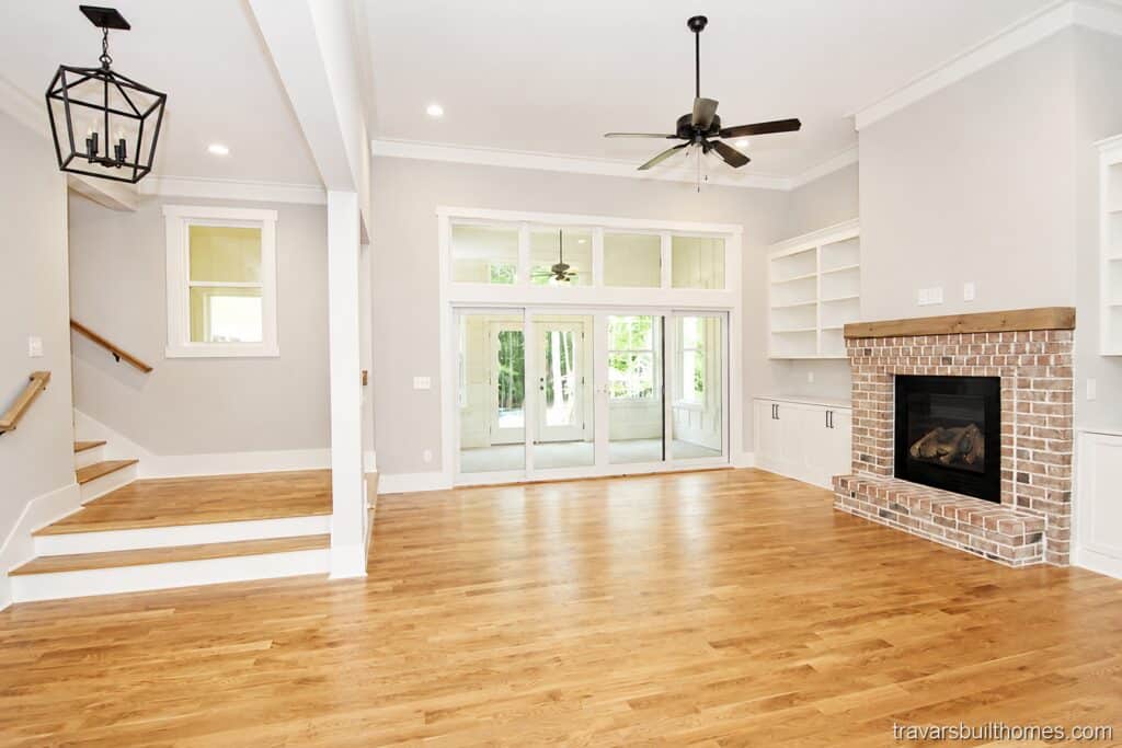 Spacious living area showcasing a beautiful red brick fireplace with a rustic wood mantel, flanked by white custom built-in bookshelves and base cabinetry, positioned next to a multi-slide glass patio door, built by Travars Built Homes.