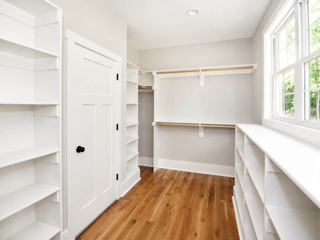 Generous master walk-in closet detailing rich hardwood floors, custom white built-in shelving arrays, wooden hanging rods, and a window overlooking the property, crafted by Travars Built Homes.