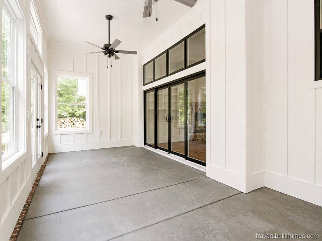 Enclosed outdoor sunroom or patio featuring a smooth concrete floor, white board-and-batten walls, a ceiling fan, and large black-framed multi-slide glass doors with transoms, built by Travars Built Homes.