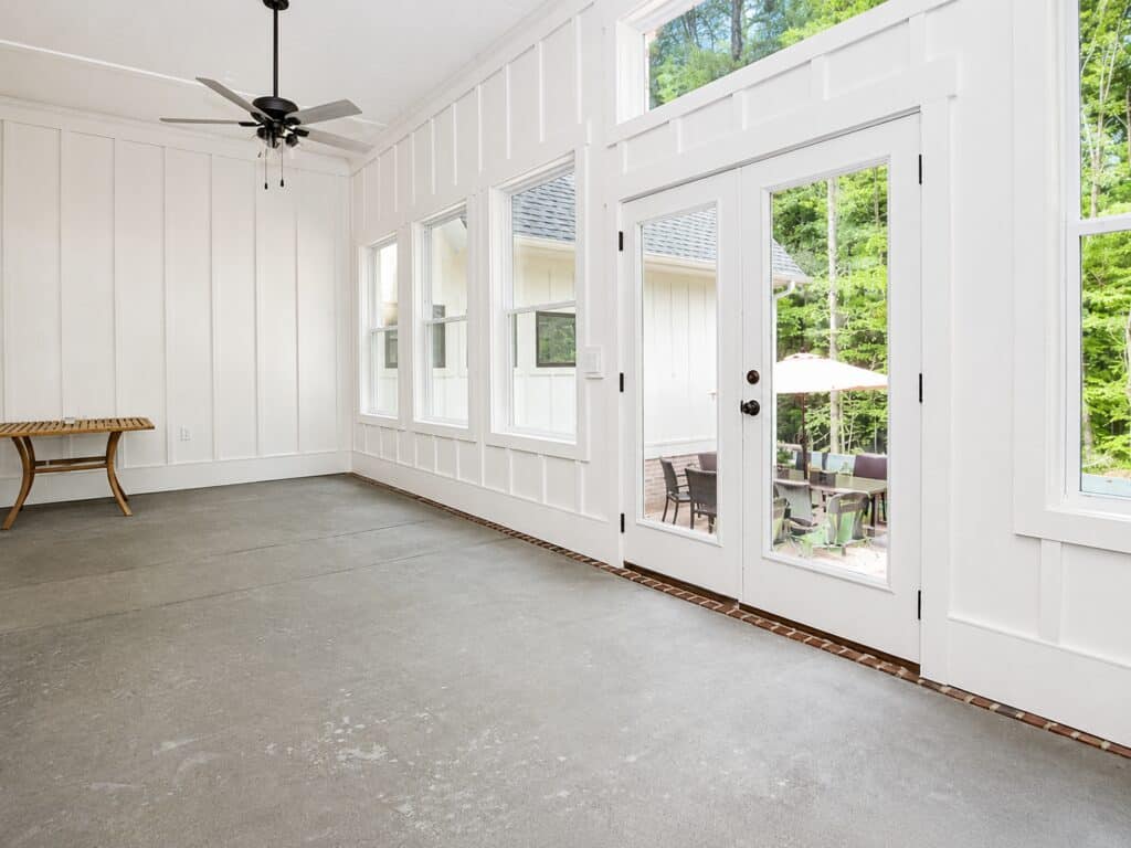 Alternative view of the enclosed concrete patio room, highlighting white vertical panel walls, a dark ceiling fan, and double glass French doors that open out to an exterior paved seating area, built by Travars Built Homes.