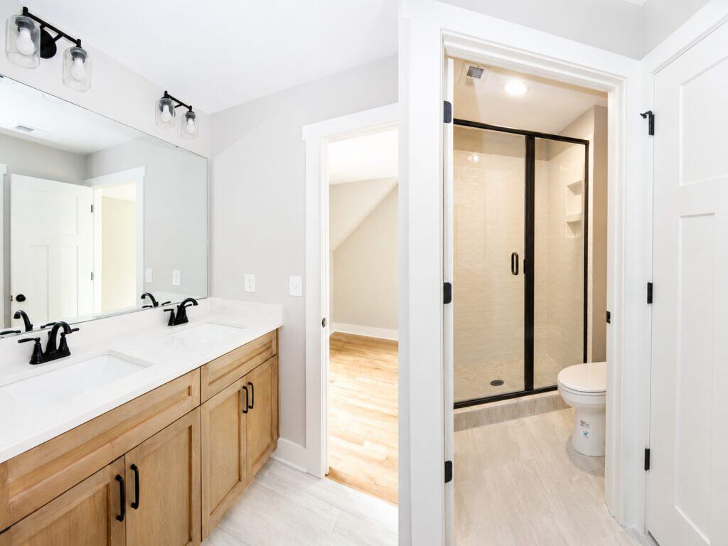 Split view of a secondary bathroom detailing a double-sink natural wood vanity with matte black faucets on the left, alongside a view toward the toilet and black-framed walk-in shower on the right, designed by Travars Built Homes.