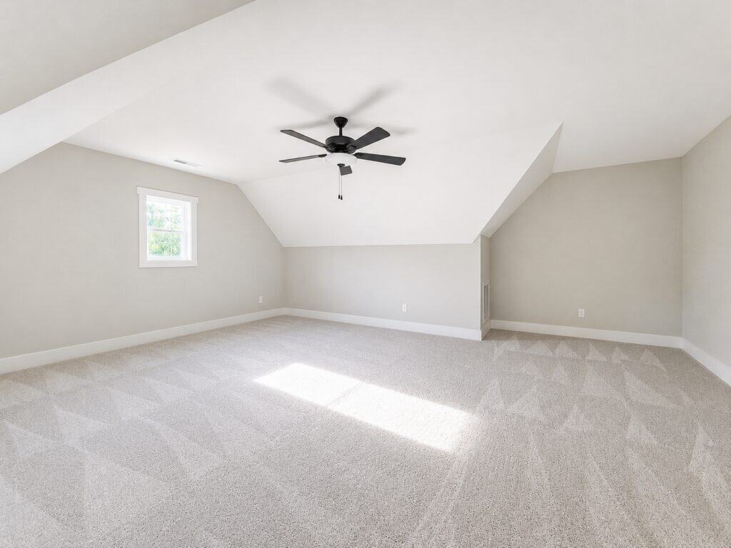 Expansive custom bonus area featuring soft patterned carpeting, light greige walls, and angled ceilings with a modern dark fan, crafted by Travars Built Homes.