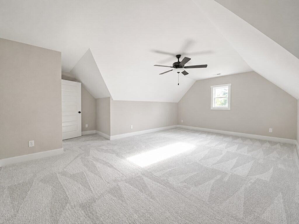 Large finished upstairs bonus room or loft space highlighting soft grey walls, sloped ceilings, plush carpeting, and a central ceiling fan, designed by Travars Built Homes.
