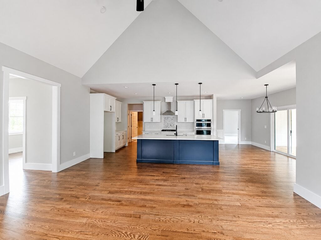 Expansive open-concept kitchen highlighting a contrasting blue island with a stainless steel sink, crisp white cabinetry, modern pendant lighting, and a clear sightline into the vaulted living area, crafted by Travars Built Homes.