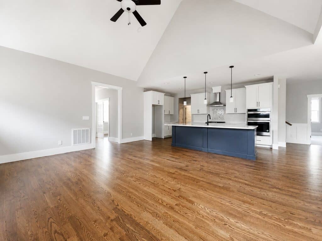 Wide view of a custom kitchen and living space featuring a deep blue island, white shaker cabinets, rich oak hardwood flooring, and a dramatic vaulted ceiling, designed by Travars Built Homes.