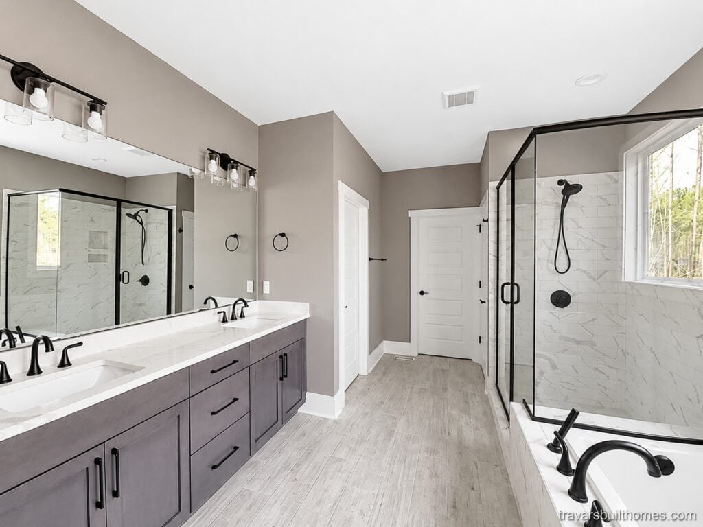 Spacious en-suite bathroom featuring a long grey double vanity with a quartz countertop, matte black fixtures, and a large wet room area combining the tub and shower behind a glass enclosure, designed by Travars Built Homes.