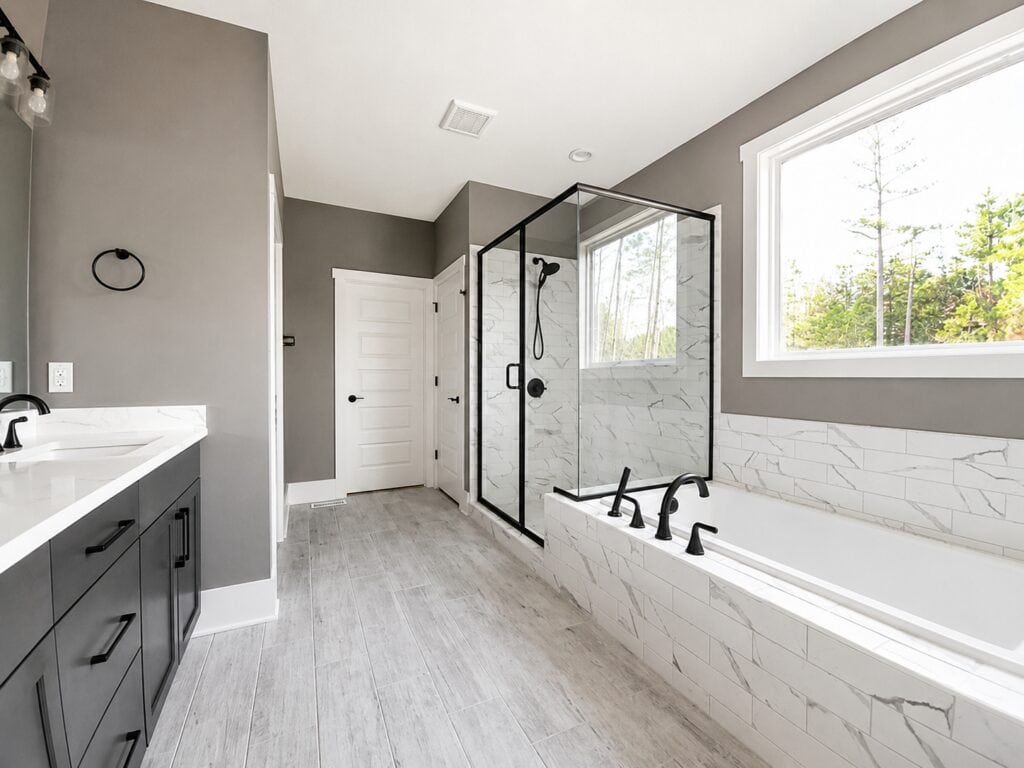 Luxurious custom bathroom highlighting a built-in soaking tub flowing into a seamless glass walk-in shower with marble-style tile, paired with a dark grey double vanity, built by Travars Built Homes.