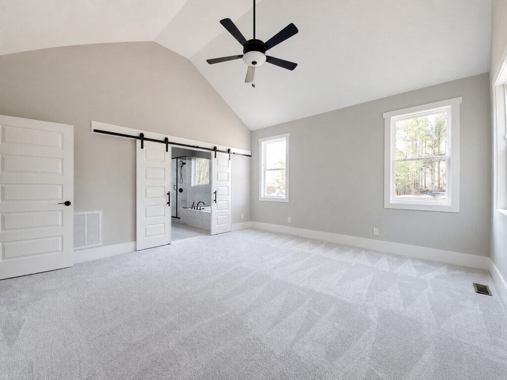 Elegant primary bedroom featuring a soaring vaulted ceiling, soft neutral tones, patterned carpeting, and sliding white double barn doors opening to the en-suite bathroom, designed by Travars Built Homes.