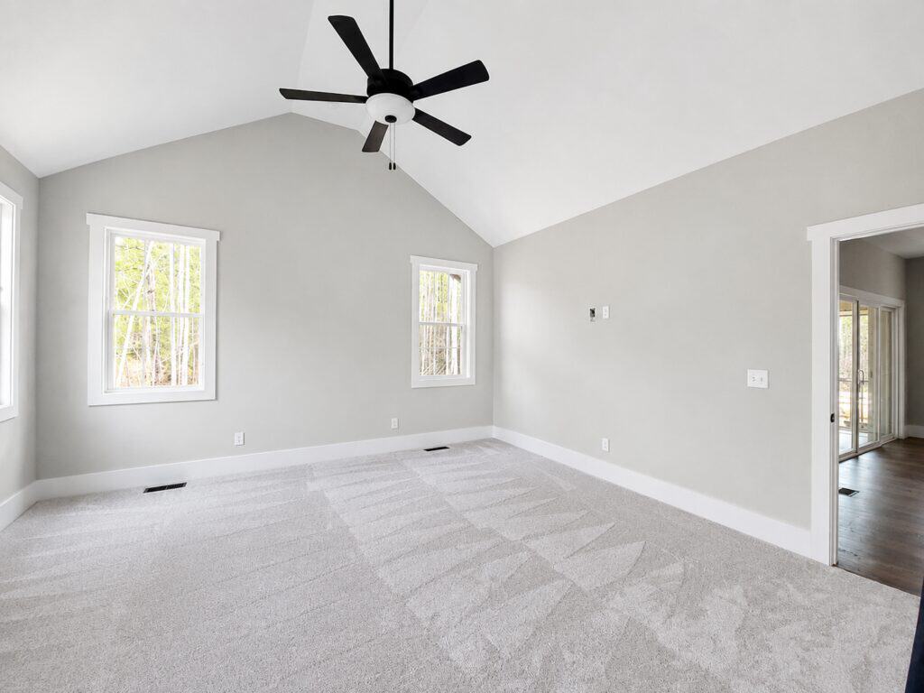 Spacious custom primary bedroom showcasing a vaulted ceiling with a modern black fan, plush grey patterned carpeting, soft grey walls, and glass doors leading to the rear porch, built by Travars Built Homes.