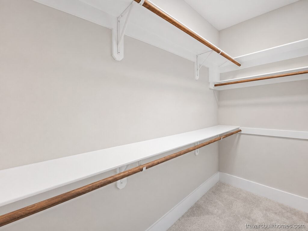 Detailed view of a custom walk-in closet highlighting built-in white laminate shelving, sturdy wooden hanging rods, and soft neutral carpeting, designed by Travars Built Homes.