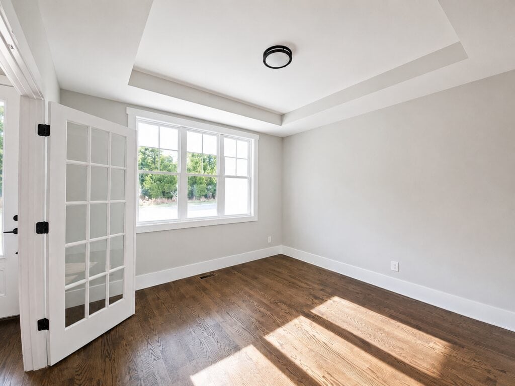 Custom flex room or study featuring white double doors with glass panes, warm oak hardwood flooring, and a large window providing ample natural light, built by Travars Built Homes.
