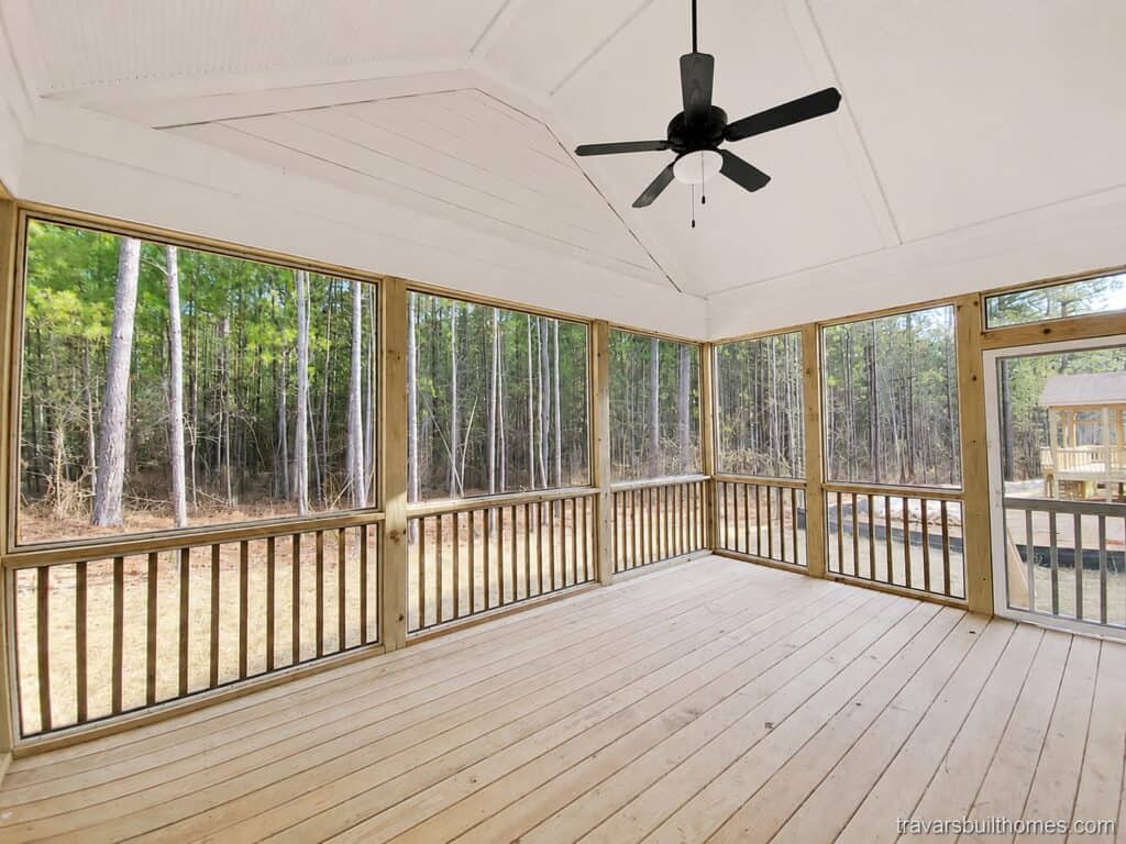 View of a spacious screened-in porch showing a dark vaulted ceiling, a black ceiling fan, natural wood framing, and multiple large screens looking out to a densely wooded area, built by Travars Built Homes.