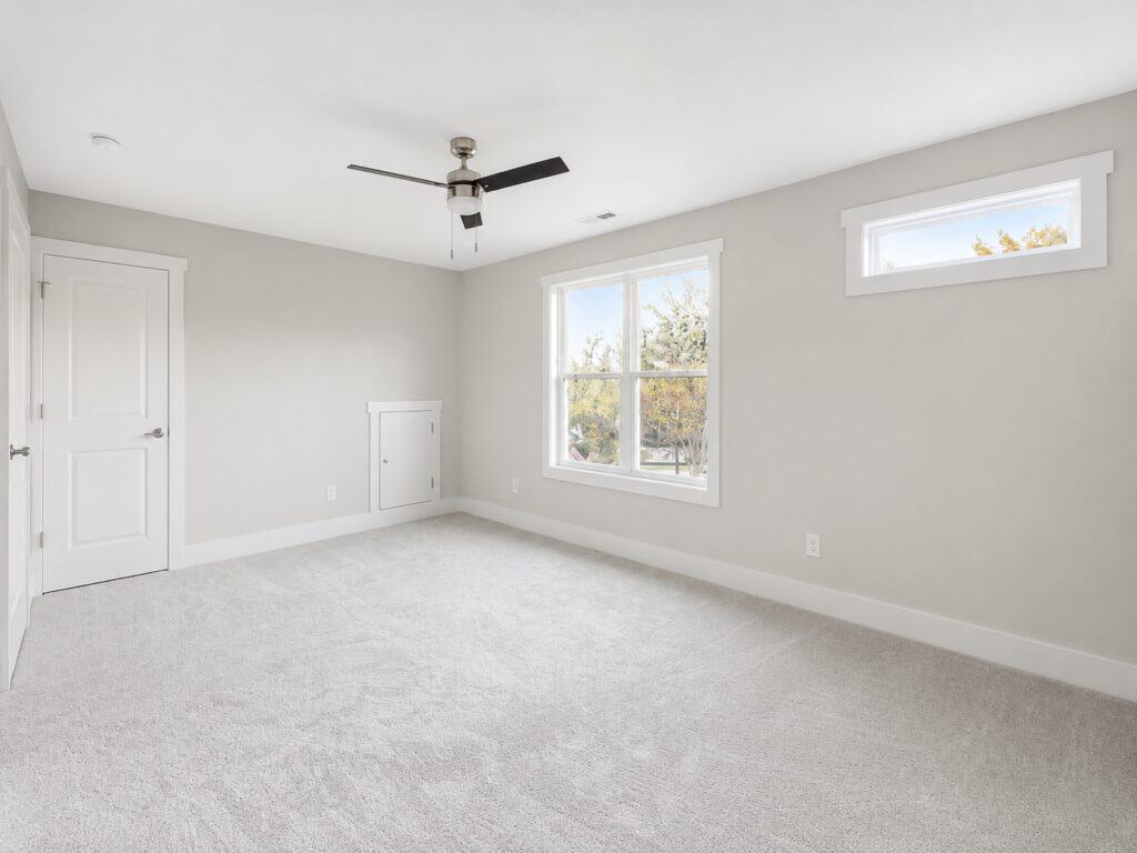 Bright custom secondary bedroom featuring light grey walls, a modern ceiling fan, soft carpeting, and a small transom window providing additional natural light, by Travars Built Homes.