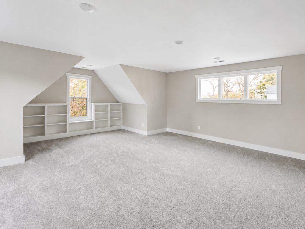 Wide view of a large upstairs loft or bonus area highlighting built-in low shelving beneath a dormer window, sloped ceilings, and neutral carpeting, designed by Travars Built Homes.