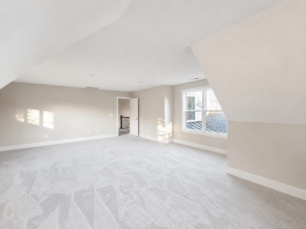 Spacious custom finished bonus room featuring sloped ceilings, soft greige walls, plush carpeting, and multiple windows providing natural light, built by Travars Built Homes.