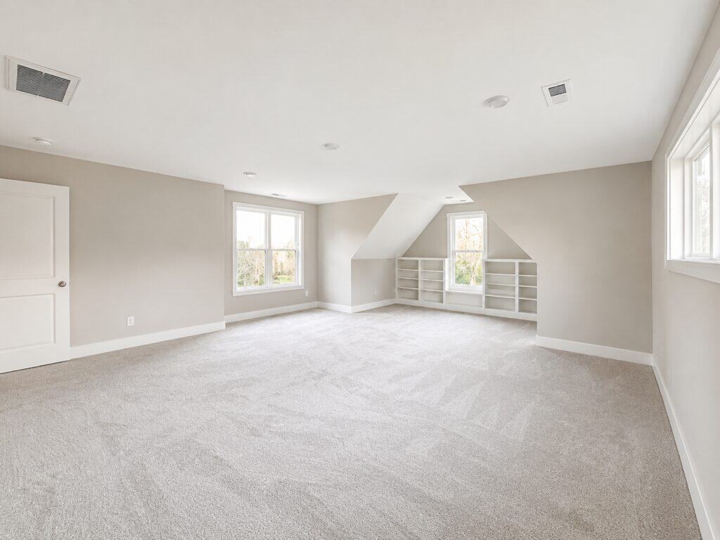 Custom finished upstairs bonus space showcasing built-in white shelving below dormer windows, plush grey carpeting, and recessed lighting, crafted by Travars Built Homes.