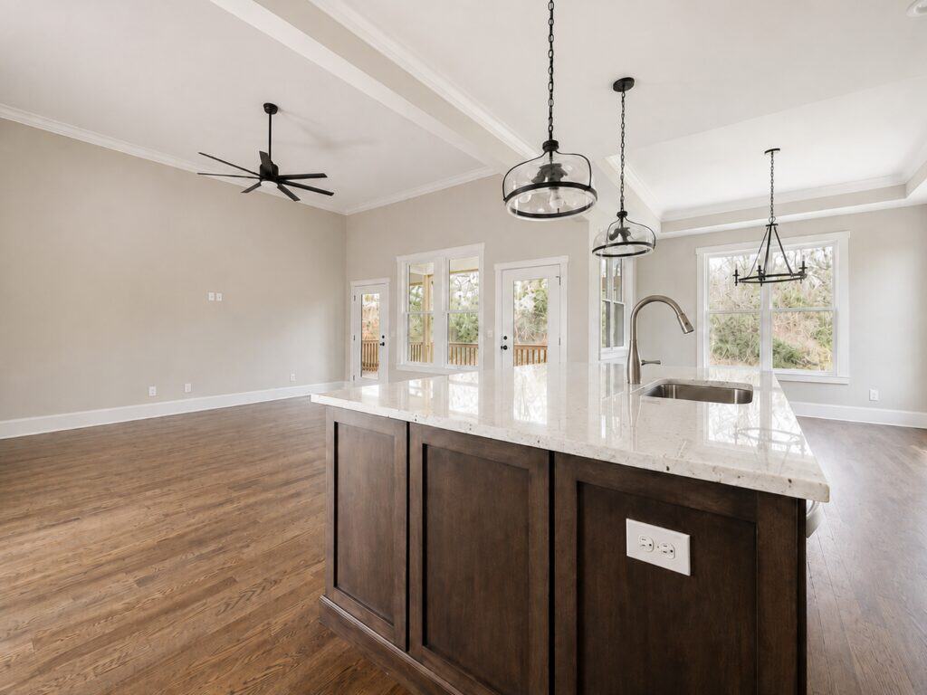 Bright open-concept kitchen and living area featuring a large dark wood island with a white quartz top, rich hardwood flooring, and multiple glass doors providing natural light and access to the outdoor living space, crafted by Travars Built Homes.
