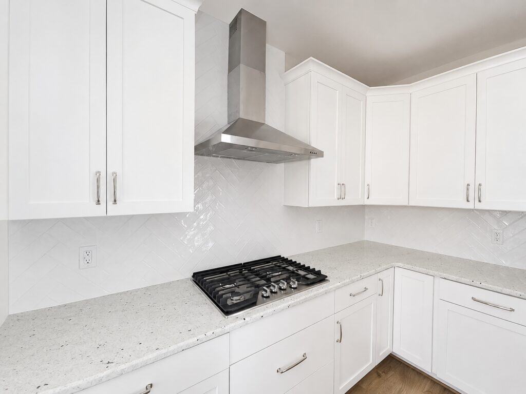 Detail view of a custom kitchen corner highlighting crisp white shaker cabinets, a sleek stainless steel gas cooktop with a matching range hood, and a glossy white herringbone tile backsplash, by Travars Built Homes.
