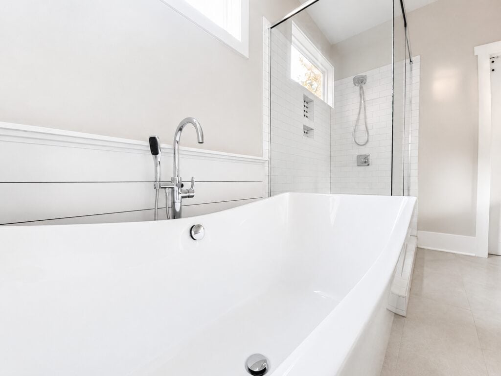 Close-up of a modern freestanding soaking tub paired with a sleek chrome floor-mounted faucet, set against crisp white shiplap wainscoting next to a glass-enclosed shower, built by Travars Built Homes.